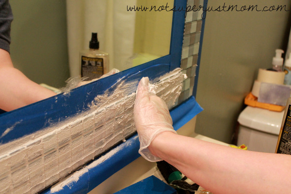 A gloved hand spreading grout across the mosaic tile border, pushing it down into the gaps between tiles